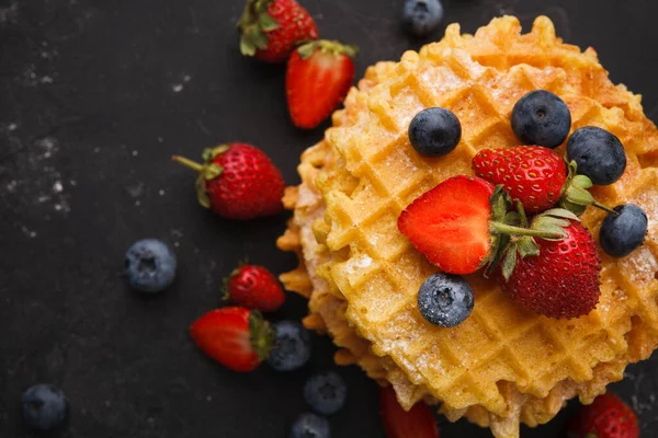 Bir yığın altın waffle karanlık bir yüzeyde duruyor. Taze çilekler ve yaban mersinleri waffle 'ların etrafına saçılırlar. Bu kurgu kahvaltı ya da tatlı için cazip bir sahne yaratır..