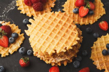 Waffle 'lar etraflarını saran çeşitli taze meyvelerle karanlık bir yüzeye yerleştirilir. Çilekler, yabanmersinleri ve ahududular etrafa saçılmış..
