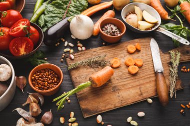 Bir grup taze sebze ahşap bir masaya yayılır. Domates, havuç ve otlar var. Yakınlarda bir bıçak, bir kase fasulye ve şifalı ot da var. Bu sahne yemek hazırlama aktivitesini gösterir.