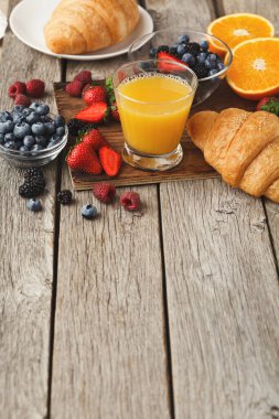 Çeşitli kahvaltılık eşyalar ahşap bir masada düzenlenir. Kruvasan, böğürtlen ve portakal gibi taze meyveler ve bir bardak meyve suyu var. Bu düzen sabah yemeği için ideal..