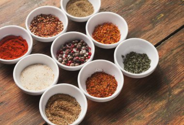 On küçük beyaz kâse seti ahşap bir yüzey üzerinde çeşitli baharatlar barındırır. Baharatlar kırmızı toz, bitkiler ve renkli tohumlar içeriyor. Her kasenin yemek pişirmek için kendine has bir çeşidi var..