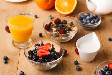 Taze meyve, yulaf ve meyve suyu ahşap bir masada düzenlenir. Bir bardak portakal suyu yabanmersini, böğürtlen, çilek, ahududu ve yoğurt kaselerinin yanında durur..