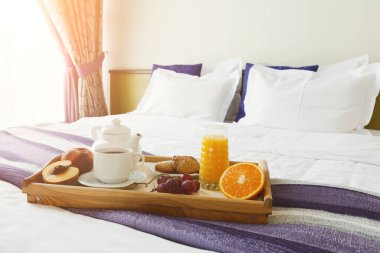 Bir tepside kahve, meyve suyu, meyve suyu ve hamur işi bir yatakta durur. Güneş pencereden parlıyor ve sıcak bir atmosfer yaratıyor. Arka planda yumuşak yatak ve renkli yastıklar görünür..