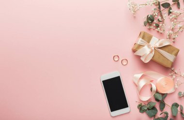 Bir kolaj kahverengi kağıda sarılı bir kurdele, bir akıllı telefon, iki nişan yüzüğü ve pembe arka planda dekoratif çiçekleri gösterir. Bu eşyalar bir kutlama ya da olay olduğunu gösteriyor..