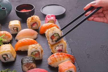 Bir el yemek çubuklarını tutar ve kara taş bir levhanın üzerine serilmiş bir koleksiyondaki suşi rulosunu toplar. Çeşitli suşi türleri yakınlarda soslarla sıralanır..