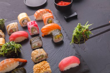 Çeşitli suşi parçalarından oluşan bir koleksiyon siyah bir taş tabakta düzenlenir. Taze balık ve sebzeler görülebilir. Bir insan yeşil garnitür almak için çubuk kullanır..