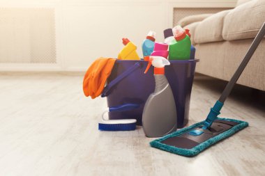 Bir temizlik malzemesi koleksiyonu yerde bir kovada düzenlenir. Yakınlarda bir paspas dururken çeşitli şişe, sünger ve eldivenler gösteriliyor. Kanepesi olan bir oturma odası..