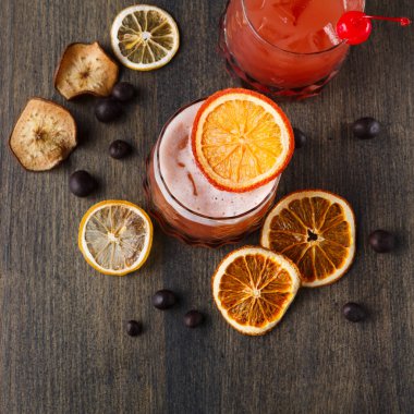 İki içki ahşap bir yüzeye oturur, biri bir dilim portakal ve kirazla süslenir. Kurutulmuş meyve dilimleri ve küçük siyah tohumlar içkilerin etrafını sarıp, olaya detay katıyor..