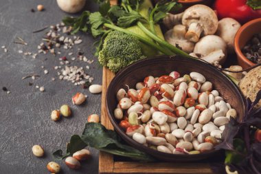 Tahta bir tahtanın üzerinde taze sebzelerle çevrili ahşap bir kasede farklı fasulye türleri sergilenir. Dekor, basit bir düzeni ve renkleri olan bir mutfak..