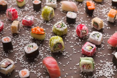 Ahşap bir yüzeyde çeşitli suşi parçaları düzenlenir. Her suşinin farklı renkleri ve malzemeleri vardır. Sushi 'nin etrafına beyaz susam tohumları saçılmış, şekillerini ve detaylarını vurguluyorlar..