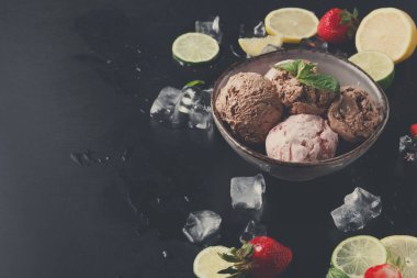 Bir kasede ortaya yerleştirilmiş üç kepçe dondurma var. Kasenin etrafında limon, limon, çilek ve buz parçaları var. Ortam karanlık ve tatlıya odaklanmamızı sağlıyor..