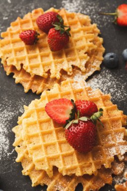 İki waffle karanlık bir yüzeye oturur. Taze çilek ve yabanmersini waffle 'ları süslüyor. Pudra şekeri tabağın tozunu alır ve sahneye biraz tatlılık katar..