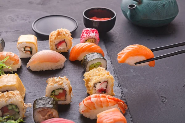 Renkli suşi parçaları siyah bir levhada sergilenir. Düzenleme çeşitli suşi türlerini içeriyor ve yemek çubukları da elinde. Yanında küçük bir kase sos var..