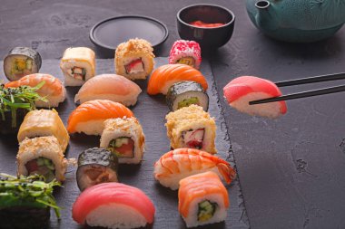 Siyah bir tabakta çeşitli suşi türleri sergilenir. Bazı parçalar karides ve yengeçle kaplıdır. Yemek çubukları bir parça toplarken yanında soslu küçük bir kase oturuyor..