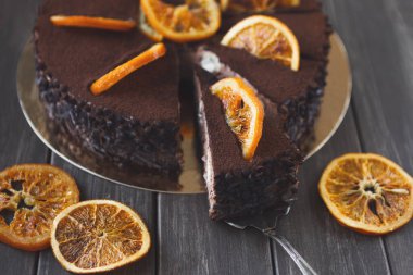 Yuvarlak bir pasta gümüş bir tepside oturur. Sergide zengin çikolata ve üzerinde portakal dilimleri yer alıyor. Pastadan bir parça kesilir, katmanlı dokusu ortaya çıkar..