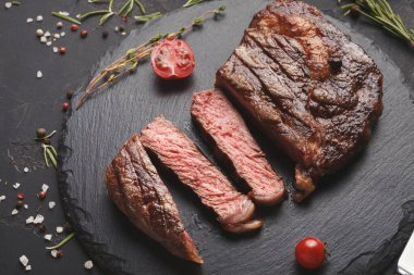 Koyu bir tahta üzerinde ızgara et dilimleri koleksiyonu düzenlenir. Etin etrafında taze bitkiler ve kiraz domatesleri var. Sahne iştah açıcı ve servis için hazır görünüyor..