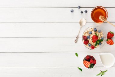 Kahvaltı malzemelerinin bir koleksiyonu beyaz ahşap bir masada düzenlenir. Çilekli ve yaban mersinli granola, bir kavanoz bal ve küçük bir kap yoğurt var. Bir de kaşık var..