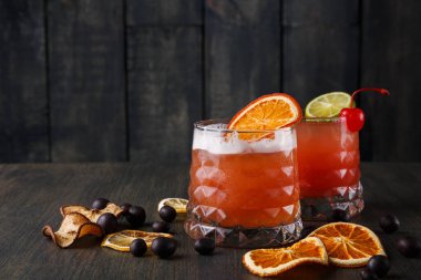 Renkli kokteyllerle dolu iki bardak koyu ahşap bir masaya yerleştirilir. Her bardağın kenarında bir dilim portakal ve limon var. Kurutulmuş meyve ve böğürtlen etrafa saçılmış..