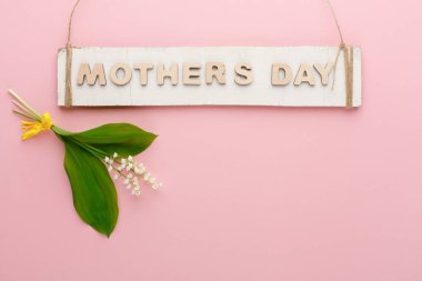 Bir tabela, Anneler Günü 'nün pembe arka planda taze çiçeklerin üzerinde asılı olduğunu gösteriyor. Çiçekler arasında sarı çiçekler ve küçük beyaz taç yaprakları da yer alıyor..