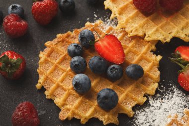 Waffle 'lar üzerine taze yaban mersini, çilek ve ahududu konmuş koyu bir yüzeyde otururlar. Etrafa pudra şekeri serpiştiriliyor, mutfakta tatlı bir sahne yaratıyor..