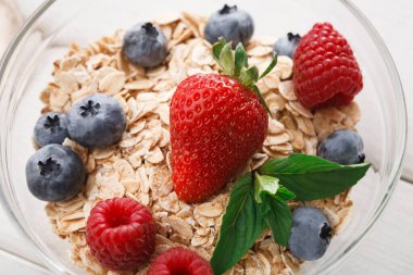 Bir kase taze çilek, ahududu, yabanmersini ve nane yapraklarıyla kaplı rulo yulaf içerir. Kase ahşap bir masaya konur, basit bir kahvaltı sahnesi sergilenir..