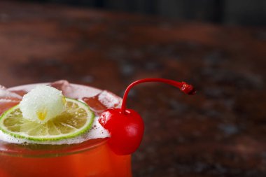 Bir kokteyl bardağı içinde buz olan bir yüzeye oturur. Bir dilim limon ve kiraz içeceği süsler. Ayar koyu bir tona sahip, içki detaylarına odaklanıyor..