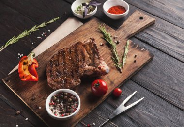 Taze ızgara biftek ahşap bir tahtanın üzerinde duruyor. Etin etrafında otlar, kiraz domatesleri ve baharatlar var. Düzenek davetkar ve yemek için hazır görünüyor..