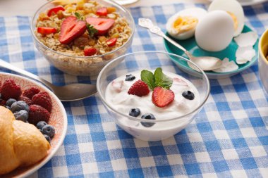 Sabah yemeği yoğurt, çilek ve ahududu ile birlikte bir kase mısır gevreği ve haşlanmış yumurta ile sergilenir. Ayarda mavi kareli masa örtüsü var..