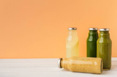 Bir masada dört meyve suyu şişesi düzenlenir. Şişeler farklı renklerde farklı içecekler içeriyor. Arka plan sade turuncu. Bu kurulum bir mutfak alanında canlı bir ekran oluşturur.