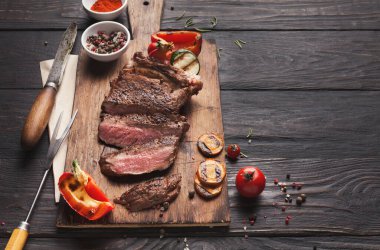 Ahşap bir tahtanın üzerinde ızgara biftek parçaları koleksiyonu. Biber ve domates gibi taze sebzeler etin yanına yerleştirilir. Baharat kaseleri arka planda durur..