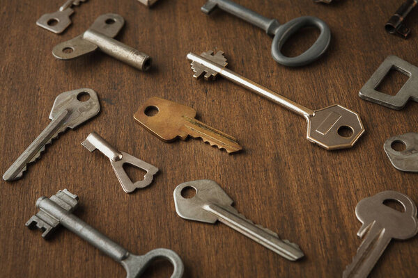 Various keys are laid out on a wooden table in a random pattern. The keys vary in size and design, showcasing their distinct shapes and functions. This collection highlights their different uses.