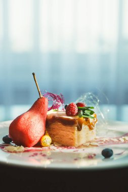 Bir tabak üzerinde bir tatlı seti düzenlenir. Üzerinde meyve ve armut olan bir parça kek var. Sahnenin arka planındaki pencereden gelen yumuşak bir ışık var..