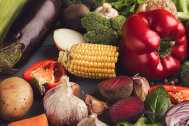 Farklı türde sebzeler karanlık bir yüzeye dizilir. Sahnede mısır, kırmızı biber, brokoli, sarımsak, patates ve diğer taze ürünler yer alıyor. Renkler ve şekiller çekici bir görünüm yaratıyor..