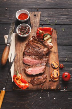 Et ızgarada pişirilir ve ahşap bir tahtada dilimlenir. Yakınlarda baharatlar ve renkli sebzeler var. Ayarlar, doğal ışık alan bir mutfak atmosferini gösteriyor..