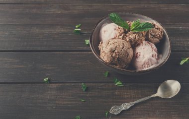 Bir kasede bir kepçe çikolata ve çilekli dondurma var. Nane yaprakları dondurmanın üzerinde oturur. Ahşap masa kırsal bir his katıyor. Gümüş bir kaşık kasenin yanında duruyor..