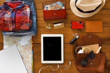 Çeşitli seyahat malzemeleri ahşap bir masanın üzerine serilmiştir. Giysiler, para, belgeler ve cihazlar gelecek bir gezi için düzenlenir. Bir harita ve tablet de mevcut.