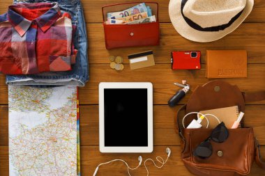 Seyahat malzemeleri ahşap bir masanın üzerine serilmiştir. Kıyafetler, içinde para olan bir cüzdan, pasaport, iPad ve harita var. Bu sahne bir gezi düzenini gösterir.
