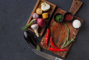 Tahta kesme tahtasında taze sebzeler ve bitkiler bulunur. Ürünler arasında mısır, patlıcan, kırmızı biber ve yeşil soğan yer alıyor. Tahtanın yanında bir bıçak duruyor..