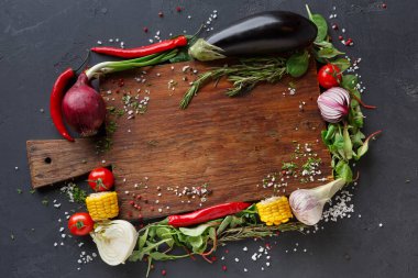Taze sebzeler ve bitkiler ahşap bir kesme tahtasının etrafına konur. Kırmızı soğan, patlıcan, domates, mısır ve çeşitli otlar tahtanın etrafını sararak, yemek hazırlamak için renkli bir görüntü oluşturuyor..