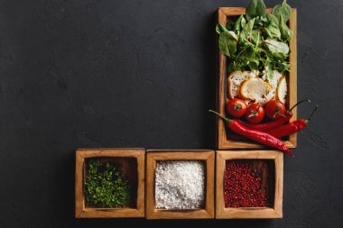 Taze bitkiler, sebzeler ve baharatlar karanlık bir mutfak yüzeyinde düzenlenir. Domatesler, biberler, ekmek ve yemek hazırlığı için hazır otlar ve baharatlar var..