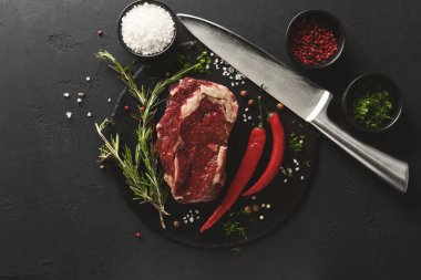 Bir parça çiğ et siyah bir kesme tahtasının üzerinde duruyor. Etin etrafında biberiye gibi otlar, küçük kaselerdeki baharatlar ve büyük bir bıçak var. Yemek hazırlığı için ortam hazır..