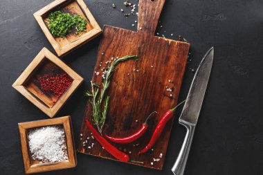 Çeşitli otlar ve baharatlar ahşap bir kesme tahtasına dizilmiştir. Taze kırmızı biber ve tuz renkli baharatlarla dolu küçük kapların yanında duruyor. Yakınlarda bir bıçak var..