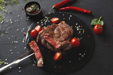 Sahne, koyu servis tabağında dilimlenmiş pişmiş bir bifteği gösteriyor. Bifteğin etrafına vişne domatesleri ve yanında küçük bir kaseye baharatlar konur..