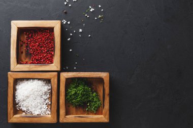 Üç ahşap kutuda farklı baharatlar ve basit bir düzende sıralanmış tuz var. Kutularda kırmızı biber, tuz ve yeşil otlar var. Bazı baharatlar karanlık bir yüzeyde kutuların etrafına saçılmış..