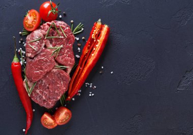 Çiğ et kesikleri taze domatesler, kırmızı biberler ve kırmızı biberlerle çevrili karanlık bir yüzeye yerleştirilir. Baharatlar ve bitkiler bir mutfak alanına tat katmak için serpilmiştir..