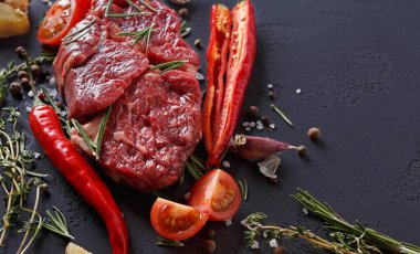 Bir parça çiğ et karanlık bir yüzeye yerleştirilir. Taze bitkiler, kırmızı biber ve domatesler etin etrafını sarar. Renkler ve dokular yemek hazırlama malzemelerini vurgular..