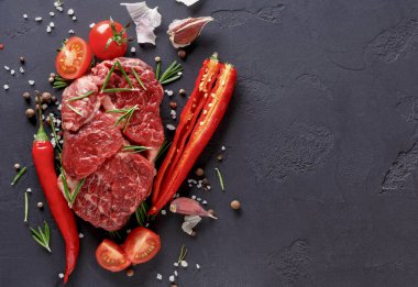 Taze çiğ et, kiraz domatesleri, sarımsak ve kırmızı biberlerin yanında koyu bir yüzeye yerleştirilir. Baharatlar ve bitkiler etrafa saçılmış, yemek pişirmek için malzemeler sergileniyor..