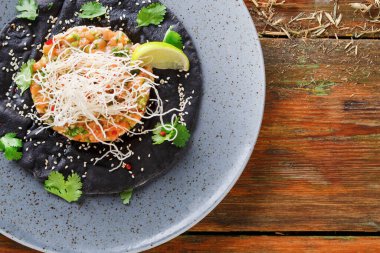 Bitki ve lahanayla kaplı siyah bir tortilla üzerinde bir deniz ürünleri tabağı sergilenir. Benekli bir tabakta, ahşap bir arka planda sunuluyor, sıradan bir yemek deneyimini yakalıyor..