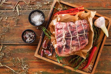 Büyük bir et parçası mutfak kağıdına sarılır ve ahşap bir tepsiye konur. Baharatlar, sarımsak ve otlar kırsal mutfak masasındaki etin etrafını sarar. Tuz yakınlardaki küçük kaselerde..