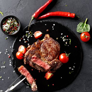 Siyah bir taş yüzeyde bir parça biftek sergilenir. Çeri domatesleri, kırmızı biberler ve bitkiler etrafına dizilmiştir. Bifteğin yanında kesmeye hazır bir bıçak var..